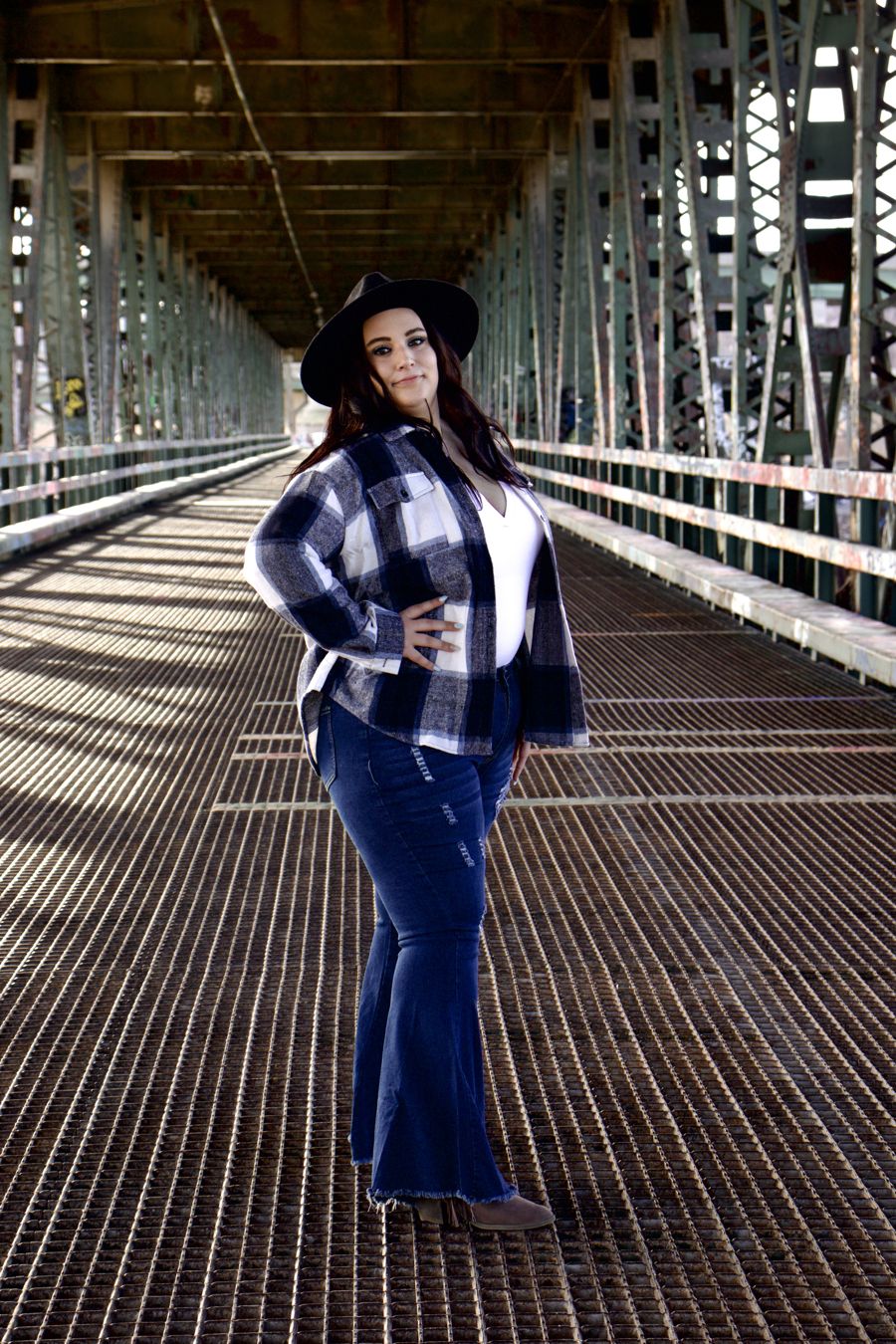 Bek standing on a steel bridge in Kansas City in a plaid jacket and wide-brim hat